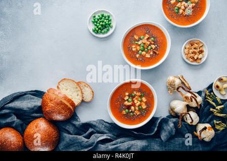 Vegan tomato soup served in three white bowls accompanied by bread rolls, roasted garlic, croutons and peas. Stock Photo