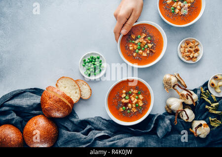 Vegan tomato soup served in three white bowls accompanied by bread rolls, roasted garlic, croutons and peas. A hand is grabbing one bowl. Stock Photo