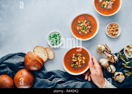 Vegan tomato soup served in three white bowls accompanied by bread rolls, roasted garlic, croutons and peas. A hand is holding one of the bowls. Stock Photo
