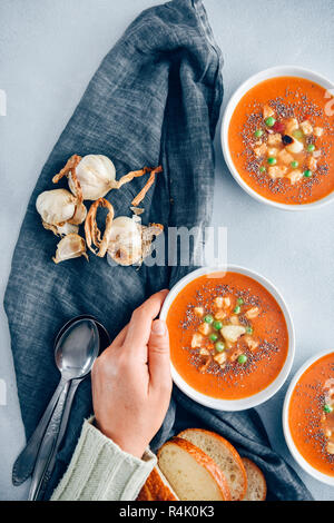 Vegan tomato soup served in three white bowls accompanied by bread rolls, roasted garlic, croutons and peas. A hand is putting one of the bowls. Stock Photo
