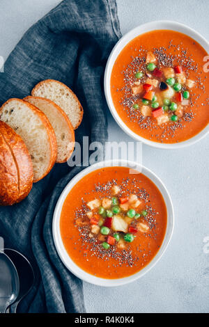 Vegan tomato soup served in two white bowls accompanied by bread rolls, roasted garlic, croutons and peas. Stock Photo