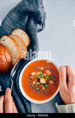 A person holding a bowl of vegan tomato soup and a spoon accompanied by bread rolls,  roasted garlic, croutons and peas. Stock Photo