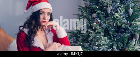 Sad girl sitting and thinking about real holiday. Serious and excited lady with a gift near the Christmas tree. copyspace for text and design. Stock Photo