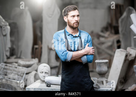 Portrait of a handsomme sculptor in blue t-shirt and apron standing in the studio with old sculptures on the background Stock Photo