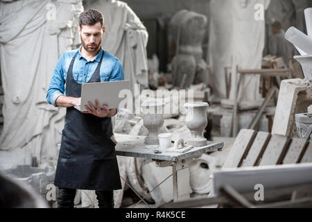 Handsome sculptor working with laptop in the atmospheric studio with old sculptures on the background Stock Photo
