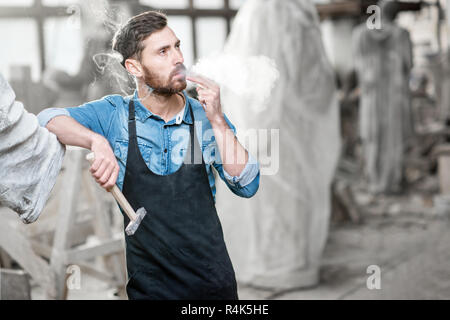 Handsome sculptor smoking a cigarette while resting after the work with sculptures in the studio Stock Photo