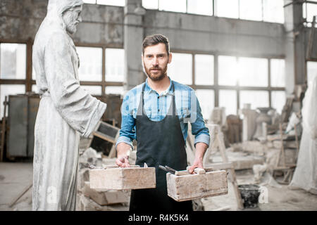 Portrait of a male sculptor in blue t-shirt and apron standing with working tools in the old studio with sculptures on the background Stock Photo
