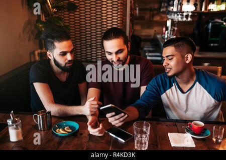 Group of mixed race young men using phone and talking in lounge bar. Multiracial friends having fun in cafe drinking coffee Stock Photo