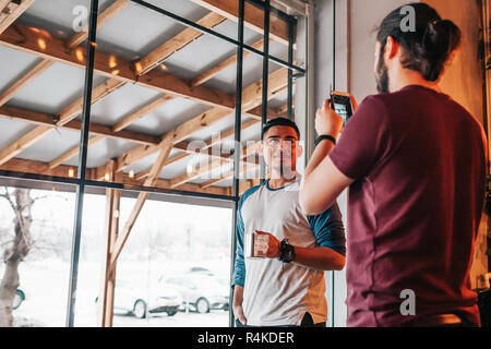 Young man takes a picture of his arabic friend on smartphone. Mixed race friends spending time in cafe. Stock Photo