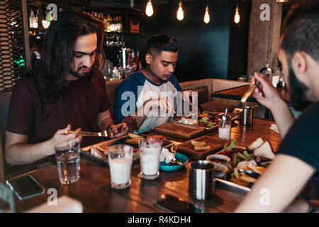 Multiracial friends eating breakfast in cafe. Young arabic men chat while having tasty food and drinks. Guys hangout together in restaurant Stock Photo