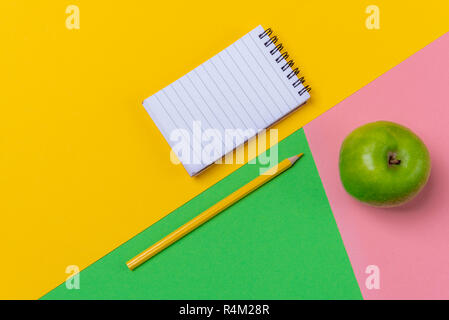 Three color table with an empty spiral notebook a yellow pencil and a green apple. Top view. Education concept. Office desk frame. Back to school. Stock Photo
