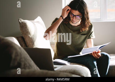 Male student studying on laptop and books sitting at home. Young man preparing for exam studying at home sitting on a couch. Stock Photo