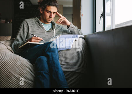 Young man sitting on a couch at home with books thinking deeply. Student making notes while studying at home with concentration. Stock Photo