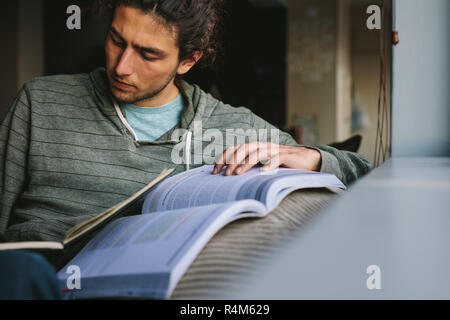 Student making notes from a reference book sitting on a couch. Young man studying seriously sitting at home. Stock Photo