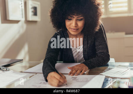 Female architect working on new building plans at her desk. African woman with curly hair making few changes in the floor plan. Stock Photo