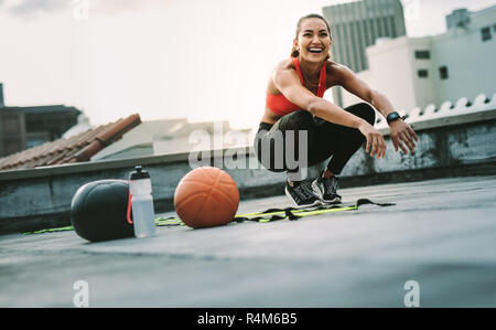Cheerful woman in fitness wear doing workout on the terrace. fitness woman doing sit ups on the rooftop with medicine ball and basketball by her side. Stock Photo