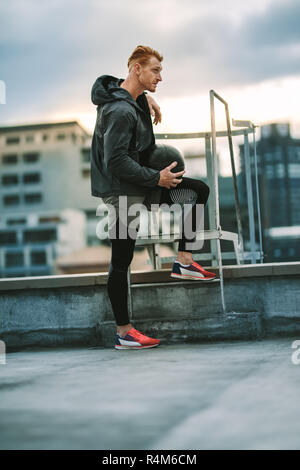 Side view of an athlete standing near a rooftop staircase and looking away. Man in fitness clothes standing relaxed holding a medicine ball on rooftop Stock Photo