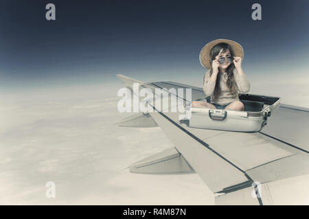 Girl sitting in the suitcase on the wing of the plane in flight Stock Photo