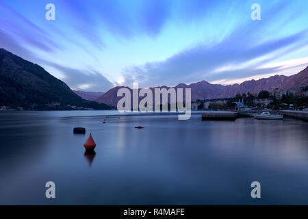 Sunset in bay of Kotor Montenegro.   Water appears flat, and wispy cloud effect, in a bay surrounded by jagged glowing mountain. Stock Photo