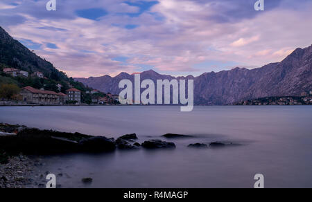 sunset in bay of Kotor Montenegro.   Water appears flat with rocks in anchor, and puffy clouds, in a bay surrounded by jagged glowing mountain Stock Photo