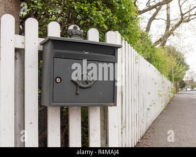 an interesting and unique black post box outside locked with an icon and attached to a white fence on a path and with hedge Stock Photo