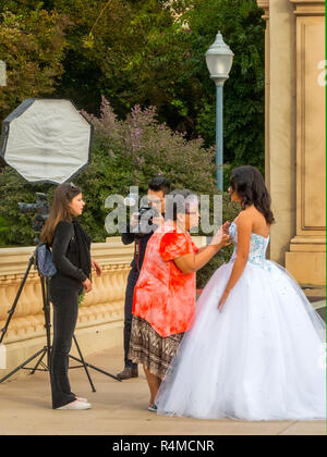 A senior Hispanic woman stylist prepares a bride in her wedding gown for formal photos by a professional photographer in Balboa Park, San Diego, CA. Stock Photo