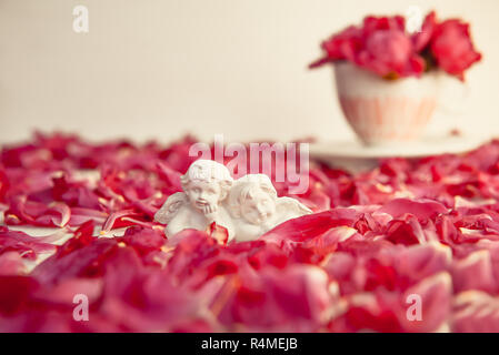 Statuette of two antique little lovely angels of the gypsum on the white background with red peony flower petals. Love, romantic, Valentines day. Vint Stock Photo