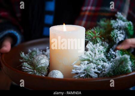 Christmas white candle in wooden tray with fire tree branches and globes, woman hands holding wooden tray with festive Christmas decorations Stock Photo