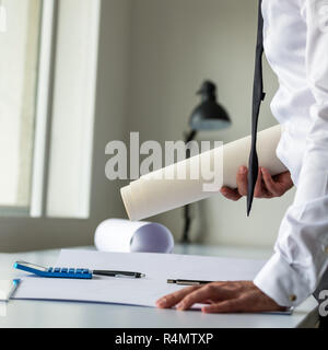 Architect or engineer leaning on his office desk holding rolled blueprints in his hands and leaning over another project on the table. Stock Photo