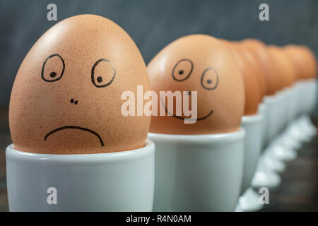 Emotions concept waiting queue or line up of ten boiled eggs in white egg cups with drawn happy and sad faces on a wooden table Stock Photo