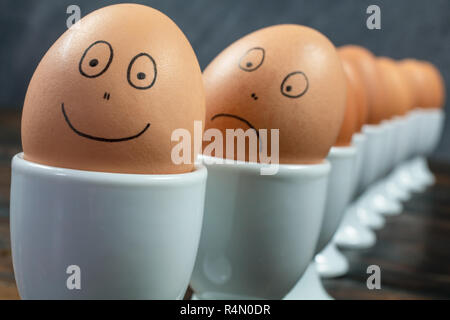 Concept waiting queue or line up of ten boiled eggs in white egg cups with drawn happy and sad faces on a wooden table Stock Photo