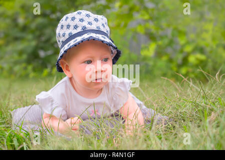 Baby girl sitting on green grass. Little princess in white dress lying on green grass in the garden. Portrait of toddler in the hat. Summer vacation b Stock Photo