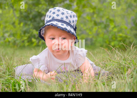 Little princess in white dress lying on green grass in the garden. Baby girl sitting on green grass. Portrait of toddler in the hat. Summer vacation b Stock Photo