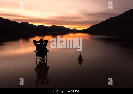 Lone figure, man, sitting on a chair relaxing, next to stone stack, as the sunsets over Loch Lomond, Scotland UK Stock Photo
