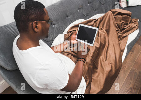 Young African American man, covered with a blanket, uses tablet. Stock Photo