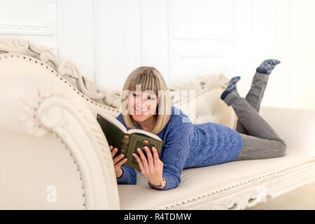 A girl in a blue dress on a white ottoman reading a book Stock Photo