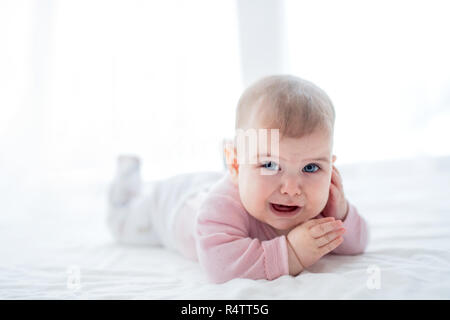 A portrait of unhappy baby girl lying on her tummy on bed indoors. Stock Photo