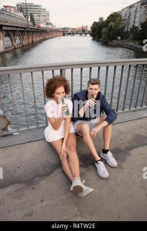 Young couple drinking beer on urban bridge over river Stock Photo