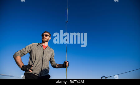 Young man on a catamaran standing and holding rope in front of a blue sky during cruising. Stock Photo