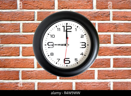 Big round clock with a black rim showing nine o'clock hangs on red brick wall front view close up Stock Photo