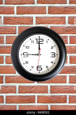 Big round clock with a black rim showing nine o'clock hangs on red brick wall front view close up Stock Photo