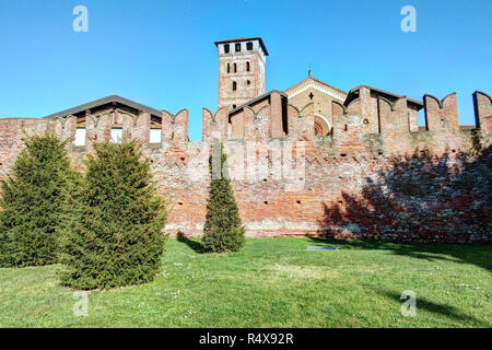 The cotto brick defense walls with Ghibelline merlons in front of a lawn with fir trees in the Saints Nazarius and Celsus abbey, Piedmont, Italy Stock Photo