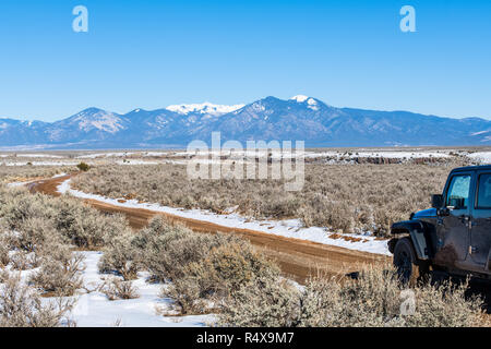 Four wheel drive vehicle on muddy road through a snow covered plain with a range of snow-capped mountains in the distance near Taos, New Mexico Stock Photo