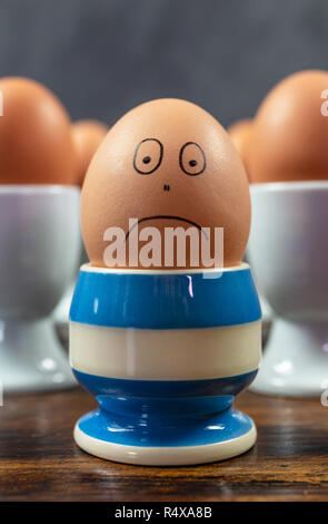 Sad emotion depression concept one individual boiled egg frowning in different blue striped egg cup surrounded by eggs in white egg cups on a wooden t Stock Photo