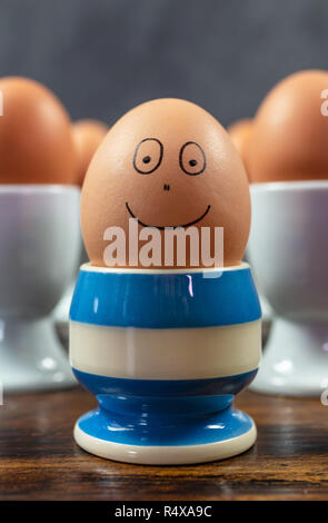 Happy emotion concept one individual boiled egg smiling in different blue striped egg cup surrounded by eggs in white egg cups on a wooden table Stock Photo