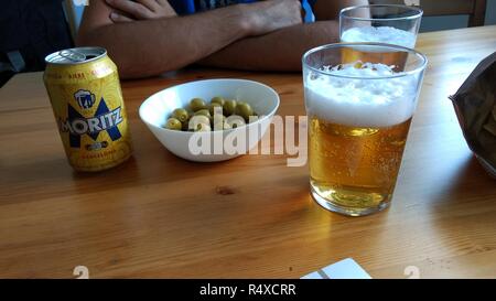 A wood color table with a typical Spanish beer can, a cup with some green olives and two glasses of lager beer in a bar Stock Photo