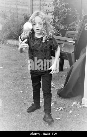 A young, Caucasian, long haired boy plays with a ball in a garden. Stock Photo