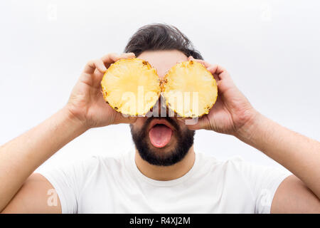 Funny and positive young man stand and cover eyes with two halfs of one pineapple. Also he show tongue on camera. Isolated on white background. Stock Photo
