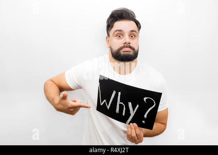 Strange young man showing emotion on face and looking at camera. He holds polate with written word Why. Also he points on it. Isolated on white background. Stock Photo