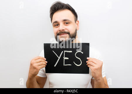 Postitive but strange young man stand and look at camera. He holds black plate with written work yes. Guy holds it with both hands. Isolated on white background. Stock Photo